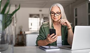 a woman sitting at a table with her laptop open and looking at her cell phone