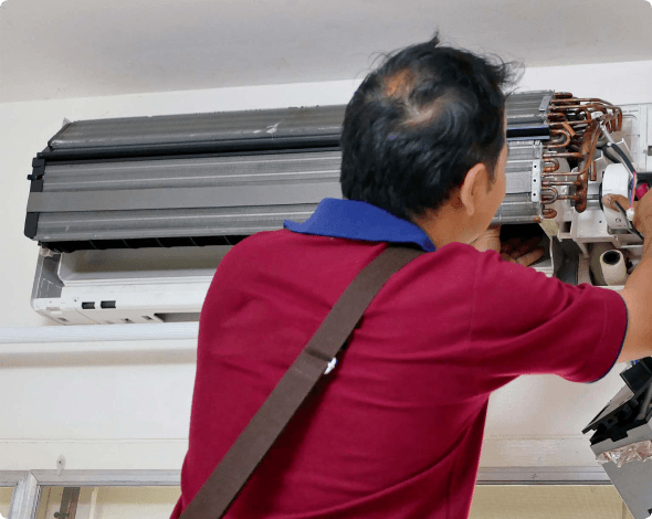 Older man in a red shirt performs repairs on a broken air conditioning unit.